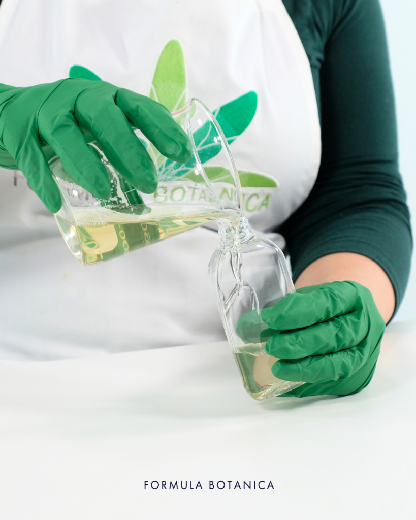 gloved hands showing step 4 of making a micellar water. Pouring finished micellar water formulation from beaker into container