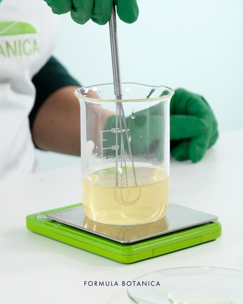 glove hands showing step 2 of how to make a micellar water. Stirring liquids in a beaker with a mini whisk
